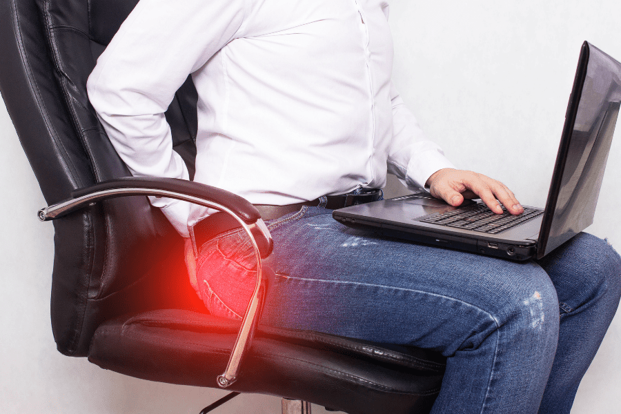 While sitting on a chair, using a laptop, a man experience pain from Hemorrhoids